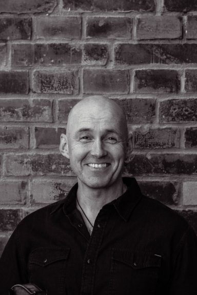 Leende man med skallat huvud framför en mur av tegelstenar.