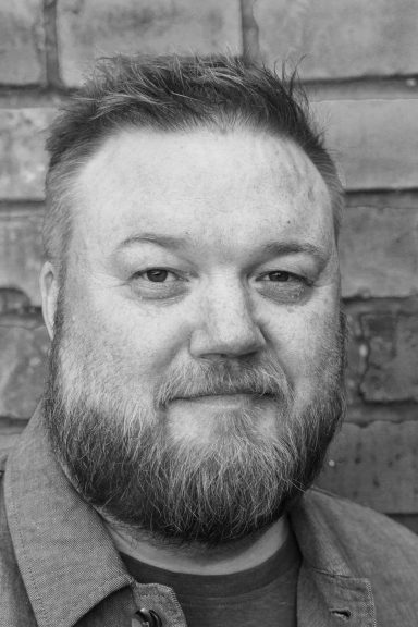 Porträtt av en man med skägg och kort hår, mot en tegelvägg.