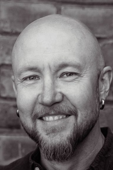 Svartvit porträtt av en skäggig man med kort hår och örhängen, leende.