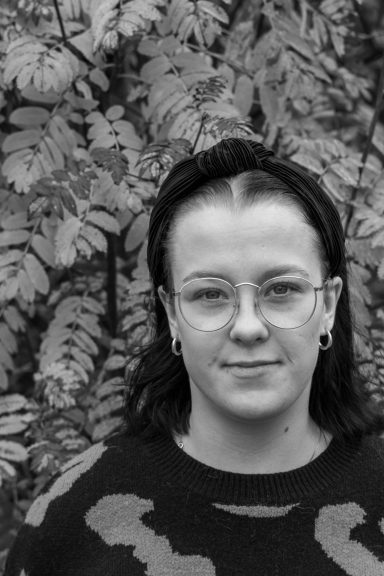 Porträtt av en person med glasögon och svart tröja, omgiven av växtlighet.