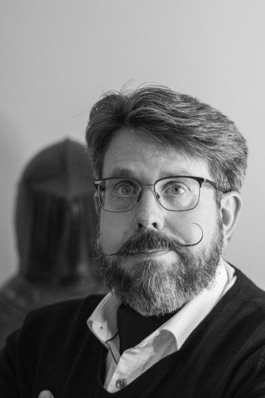 En man med glasögon och mustasch står framför en skulptur av en riddare.