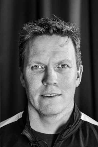 Porträtt av man med kort hår och neutral ansiktsuttryck, svart bakgrund.