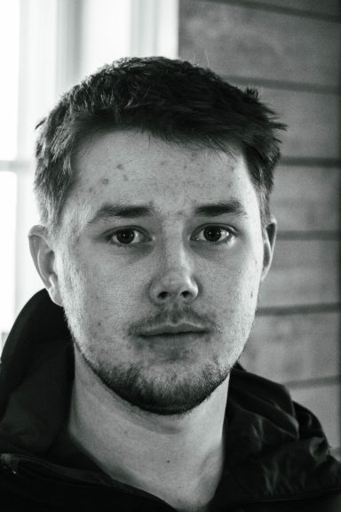 Porträtt av en ung man med kort hår och skägg, svart jacka och neutral ansiktsuttryck.