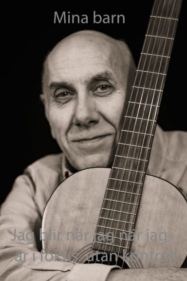 En äldre man med gitarr, ler mot kameran med en mjuk bakgrund.